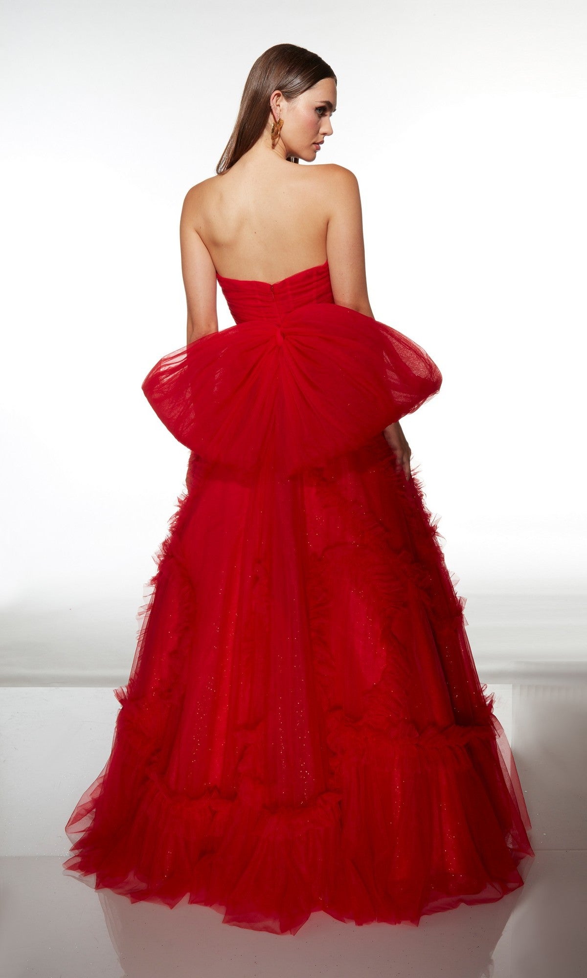 A woman stands with her back to the camera in the Alyce 62119 long prom dress, featuring a strapless corset bodice and a large tulle bow detail on the back of the floor-length red gown.